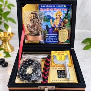 May include: A black velvet-lined box containing a Shani Dev idol, a prayer bead necklace, a metallic Shani Yantra plate, a silver-toned trident-shaped tool, and a printed card with Hindi text and an illustration of a deity riding a crow.