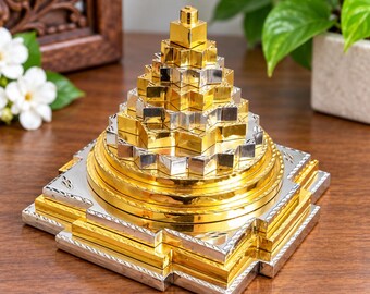 May include: A three-dimensional Sri Yantra pyramid sculpture featuring a stepped, multi-layered design. The object is constructed from alternating gold and silver-toned metallic plates, forming a geometric, pointed structure with a square base.