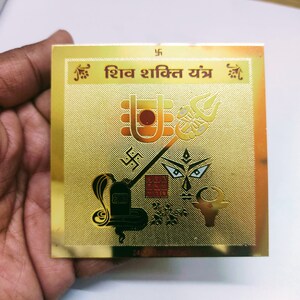 Puede incluir: Una placa cuadrada de color dorado con el texto "Shiv Shakti Yantra" en escritura hindi. La placa presenta varios símbolos religiosos hindúes, incluyendo un tridente, una serpiente y un toro.