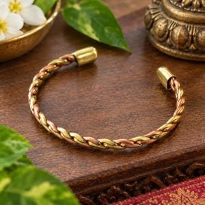 May include: A cuff bracelet featuring a twisted design made of alternating strands of polished copper and brass. The open-ended bangle has smooth, rounded metal caps at each terminal end, creating a continuous braided appearance throughout the band.