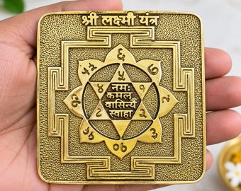 Puede incluir: Una placa cuadrada de latón Shri Lakshmi Yantra con intrincados patrones geométricos en relieve, una estrella central y texto en sánscrito. La superficie tiene un fondo texturizado con detalles pulidos en relieve, incluyendo la inscripción Shri Lakshmi Yantra en la parte superior y texto de mantra central en escritura devanagari.