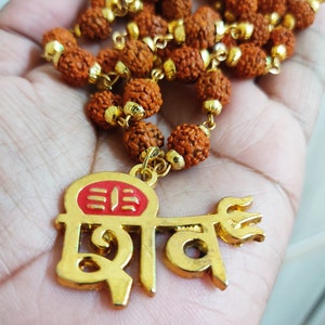 Puede incluir: Un collar con cuentas marrones texturizadas y detalles dorados. El colgante dorado, con detalles rojos, presenta la palabra "Shiva" en una escritura estilizada y un símbolo de tridente.