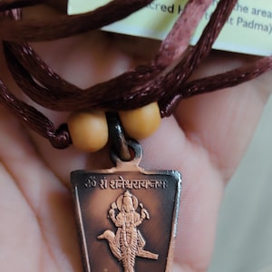 May include: A copper pendant featuring a deity riding a bird. The pendant is inscribed with the text "ॐ श्री शनिश्चराय नमः".