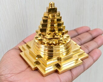 May include: A gold-colored, pyramid-shaped object with a stepped, multi-tiered design. The structure features a square base that transitions into a series of stacked, geometric layers, culminating in a small, pointed top. The surface is reflective.