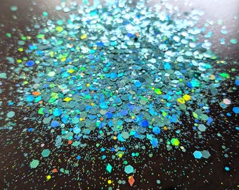 May include: A close-up of iridescent blue, green, and yellow glitter. The glitter is scattered on a dark surface.