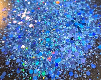 May include: A close-up of blue glitter with iridescent sparkle. The glitter is scattered on a dark surface.