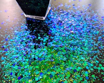 May include: A close-up of a container filled with iridescent green, blue, and gold glitter. The glitter is scattered on a dark surface.