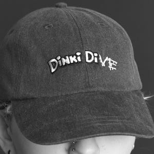 May include: A charcoal gray baseball cap with a curved brim and a six-panel construction. The front of the cap features the embroidered text Dinki Dive in white, stylized lettering with a slight arch across the center of the crown.