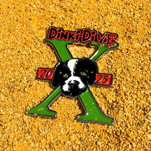 May include: A pin with the text "Dinki-Dive" in red and green, featuring a black and white dog face at the center of a green "X" shape. The pin also has the numbers "20" and "25" in red. The pin is set against a textured, tan background.