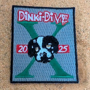 May include: Embroidered patch with the text "Dinki-Dive" in red and white. The patch features a black and white dog's face in the center, with a green "X" shape behind it. The numbers "20" and "25" are in red boxes.