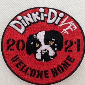 May include: Red circular embroidered patch with the text "Dinki-Di" in white, a black and white dog head, and the text "Welcome Home" in black. The year "2021" is also embroidered in white.