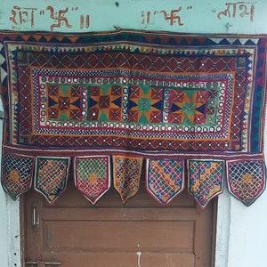 Puede incluir: Una puerta de madera marrón con una tela decorativa colgada encima. La tela tiene un patrón geométrico colorido y está adornada con pequeños espejos. El texto "श्रद्धा" "ईश्वर" "माथ" está escrito sobre la tela.