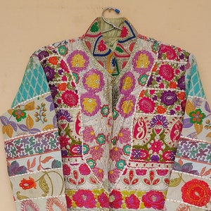 Könnte beinhalten: Eine lange, weiße Jacke mit Patchwork-Design, das bunte Blumenmuster und Stickereien zeigt. Die Jacke hat einen Stehkragen und einen Knopfverschluss.