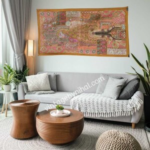 Peut inclure: Une tapisserie colorée en patchwork avec des broderies dorées est accrochée au mur au-dessus d'un canapé gris avec des coussins et une couverture. Le canapé est dans un salon avec une table basse en bois et un pouf rond.