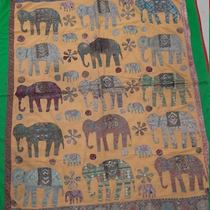 May include: A yellow fabric wall hanging with a green border features a repeating pattern of embroidered elephants in various colours and designs. The elephants are surrounded by small circles and flowers.