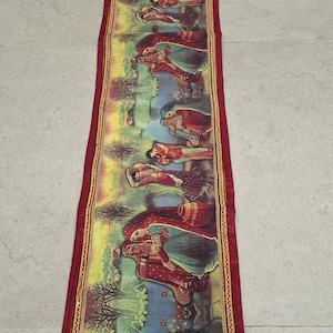 May include: A long, narrow tapestry with a maroon border and a colorful scene of people in traditional clothing. The scene is set in a lush green landscape with trees and flowers. The tapestry is decorated with gold thread embroidery.