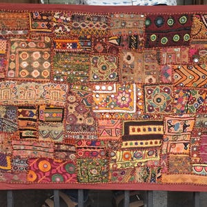 May include: A rectangular wall hanging featuring a patchwork of multicoloured fabric squares with intricate embroidery, small circular mirrors, and diverse geometric patterns. The textile is framed by a solid dark red border and includes the text vintagebynihal.