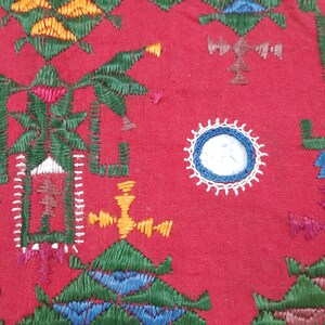 Pu&ograve; includere: Primo piano di un tessuto rosso con ricami intricati. Il design presenta forme geometriche, motivi floreali e un elemento circolare con un centro bianco e blu. Il ricamo utilizza fili verdi, gialli, blu e rosa.