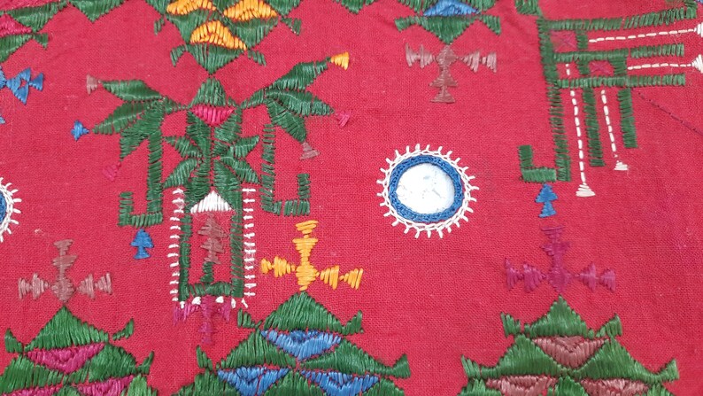 Pu&ograve; includere: Primo piano di un tessuto rosso con ricami intricati. Il design presenta forme geometriche, motivi floreali e un elemento circolare con un centro bianco e blu. Il ricamo utilizza fili verdi, gialli, blu e rosa.