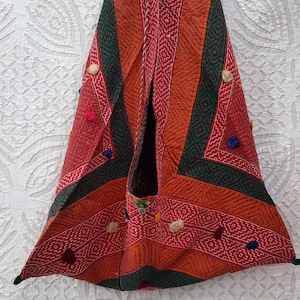 May include: A triangular-shaped, hand-stitched tote bag with a red, orange, and green geometric pattern. The bag has a unique design with a small pocket on the front and is decorated with colorful pom-poms.