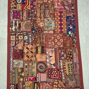 May include: A rectangular wall hanging featuring a patchwork of various fabric scraps in diverse colors and patterns. The piece includes small circular mirrors, intricate embroidery, and decorative stitching, finished with a solid red border and tassels.