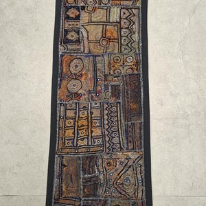 May include: Rectangular textile with a patchwork design in shades of brown, orange, and blue, framed by a black border. The textile features geometric and abstract patterns. The label says "vintagebynihal".