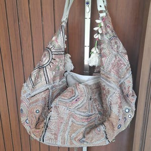 Può includere: Una borsa a spalla stile hobo con ricami complessi in tonalità beige, oro e rame. La borsa è decorata con piccoli specchi circolari, nappe bianche e dettagli a conchiglia lungo la tracolla. Include una chiusura a zip bianca.
