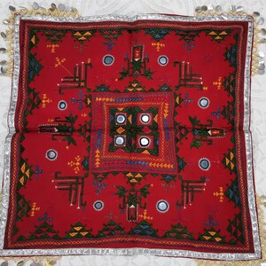 Pu&ograve; includere: Tessuto quadrato rosso con ricami intricati in verde, giallo, blu e arancione. Il design presenta motivi geometrici, piccoli specchi e un bordo argentato con decorazioni metalliche pendenti. Probabilmente un elemento decorativo o una fodera per cuscino.