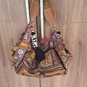 Può includere: Una borsa a tracolla stile hobo in tessuto multicolore con ricami complessi, piccoli specchi circolari e motivi geometrici. La borsa include un cinturino in pelle color cuoio e nappe nere con perline di legno. Il testo vintagebynihal è visibile.