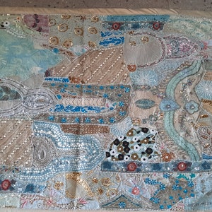 May include: A rectangular textile with a patchwork design. The piece features various shapes and patterns in shades of blue, beige, and silver, embellished with sequins and beads. The edges are finished with a cream-colored border.