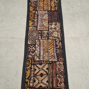 May include: A long, rectangular tapestry with a black border and a patchwork design. The tapestry is made of different fabrics and is decorated with intricate embroidery in gold, brown, and other colors.