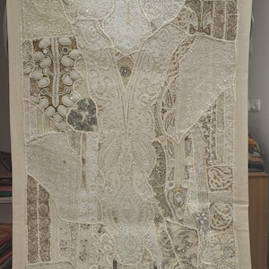 May include: Cream-colored textile wall hanging with a patchwork design. The rectangular piece features various patterns, including floral and geometric shapes, embellished with sequins and beads. The edges are trimmed with a neutral-toned border. The bottom right corner has a tassel.
