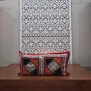 May include: A red and white patterned cushion with black and orange accents sits on a wooden platform in front of a white decorative screen.