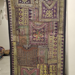 Puede incluir: Textil rectangular y ornamentado con borde negro. El tapiz presenta un diseño de patchwork con varias formas geométricas, motivos florales y bordados intrincados en tonos morados, dorados, verdes y rojos. El textil es sostenido por una persona.