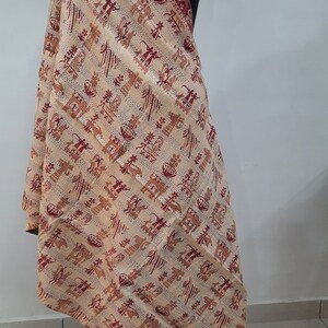 May include: A beige shawl with a red and orange pattern of stylized figures and trees. The shawl is draped over a mannequin.
