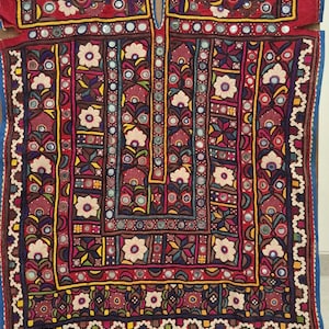 May include: A vibrantly embroidered textile with a red base, featuring intricate floral and geometric patterns in various colors. The design includes mirrors and detailed stitching, creating a rich, textured appearance. The textile is displayed on a wooden surface.
