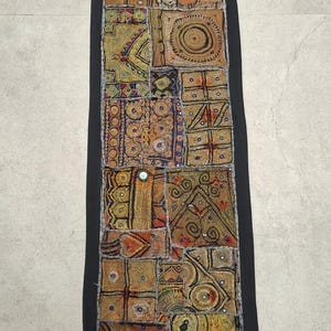 May include: A rectangular, patchwork textile with a black border. The design features various geometric patterns in shades of gold, orange, and brown. The textile has a label that says "Vintage by Nihal".