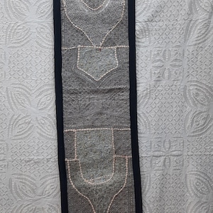 May include: A gray and white embroidered wall hanging with a black border and tassels. The embroidery features intricate patterns and designs.