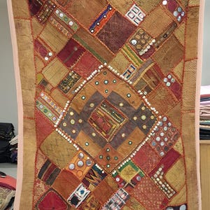 May include: A patchwork textile with a variety of warm colors, including shades of brown, orange, and red. The design features geometric shapes and mirrored accents. The textile is rectangular and appears to be a wall hanging or decorative piece.