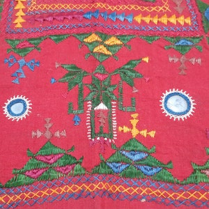 Pu&ograve; includere: Primo piano di un tessuto rosso con ricami intricati. Il design presenta forme geometriche, croci e motivi floreali in vari colori, tra cui verde, giallo, blu e rosa. Il ricamo &egrave; dettagliato e copre una parte significativa del tessuto.