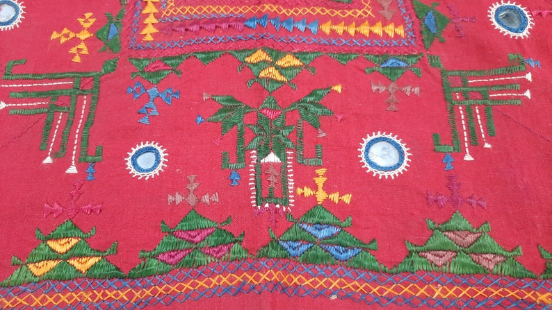 Pu&ograve; includere: Primo piano di un tessuto rosso con ricami intricati. Il design presenta forme geometriche, croci e motivi floreali in vari colori, tra cui verde, giallo, blu e rosa. Il ricamo &egrave; dettagliato e copre una parte significativa del tessuto.