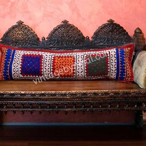 May include: A red, white, and blue embroidered pillow with geometric patterns sits on a carved wooden bench. The pillow is decorated with colorful squares and intricate details.