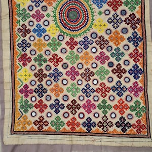 Op de afbeelding: Een kleurrijk textiel met ingewikkelde borduursels en spiegelaccenten. Het ontwerp toont een centraal cirkelvormig motief, omgeven door herhaalde kruisachtige patronen in verschillende kleuren, waaronder rood, groen, blauw en oranje. De randen zijn afgezet met een gedetailleerd patroon.