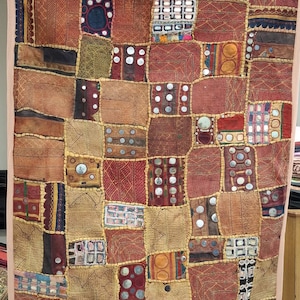 May include: A patchwork textile with a variety of textures and colors, including reds, browns, and golds. The textile is made up of numerous squares and rectangles, some adorned with small, round, reflective embellishments. The edges are trimmed with a light pink border.