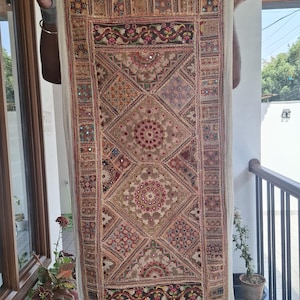 May include: A rectangular, beige textile with detailed embroidery and mirror embellishments. The design incorporates geometric patterns, floral motifs, and a central medallion. The textile is held up, displaying its intricate craftsmanship and rich colour palette.