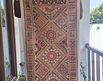 May include: A rectangular, beige textile with intricate embroidery and mirror accents. The design features geometric patterns, floral motifs, and a central medallion. The textile is held up, showcasing its detailed craftsmanship and rich color palette.