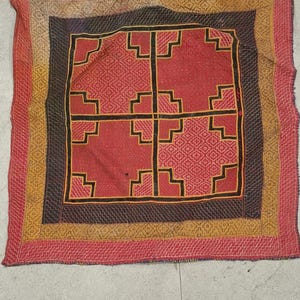 May include: A square textile with a geometric design in red, black and gold. The textile has a border with a repeating pattern.