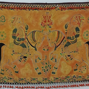 May include: A hand-painted textile wall hanging with a yellow background. Two figures are riding horses, with floral designs and a deity figure in the center. The textile has a beaded border with red tassels.