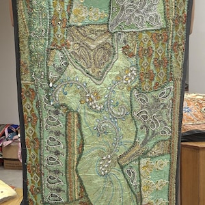 May include: A rectangular wall hanging featuring a light green fabric base with intricate patchwork, metallic embroidery, silver and gold beadwork, and sequins. The textile displays various floral and paisley patterns with a dark grey border.