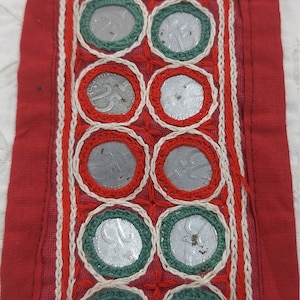May include: A red fabric strip with white and red stitching, decorated with rows of silver coins sewn into the fabric. The coins are surrounded by green and red embroidery.